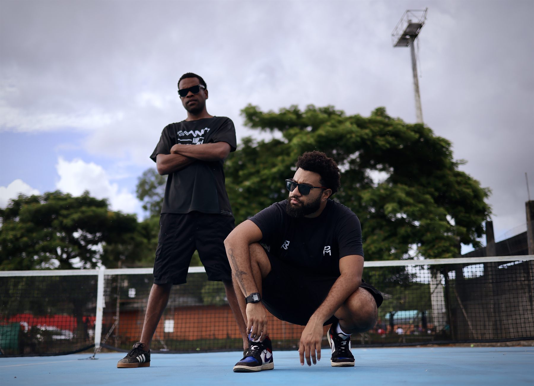 Low-angle shot on the court: a bearded artist crouches in front adjusting his sneaker while the other stands tall behind him with arms crossed against a stormy sky.