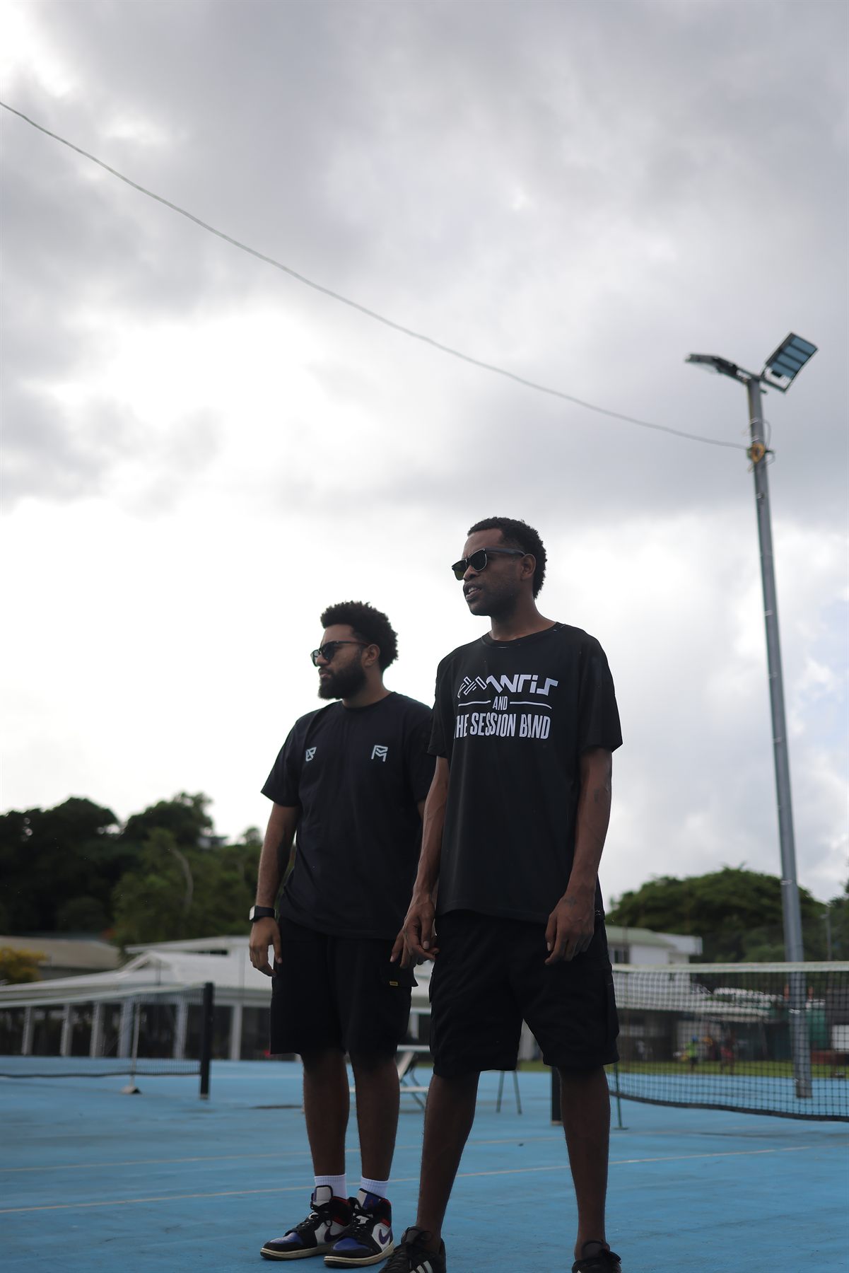 The duo standing on the blue court looking off camera, one in a Mantis and the Session Band tee, with a floodlight pole rising into heavy grey clouds behind them.