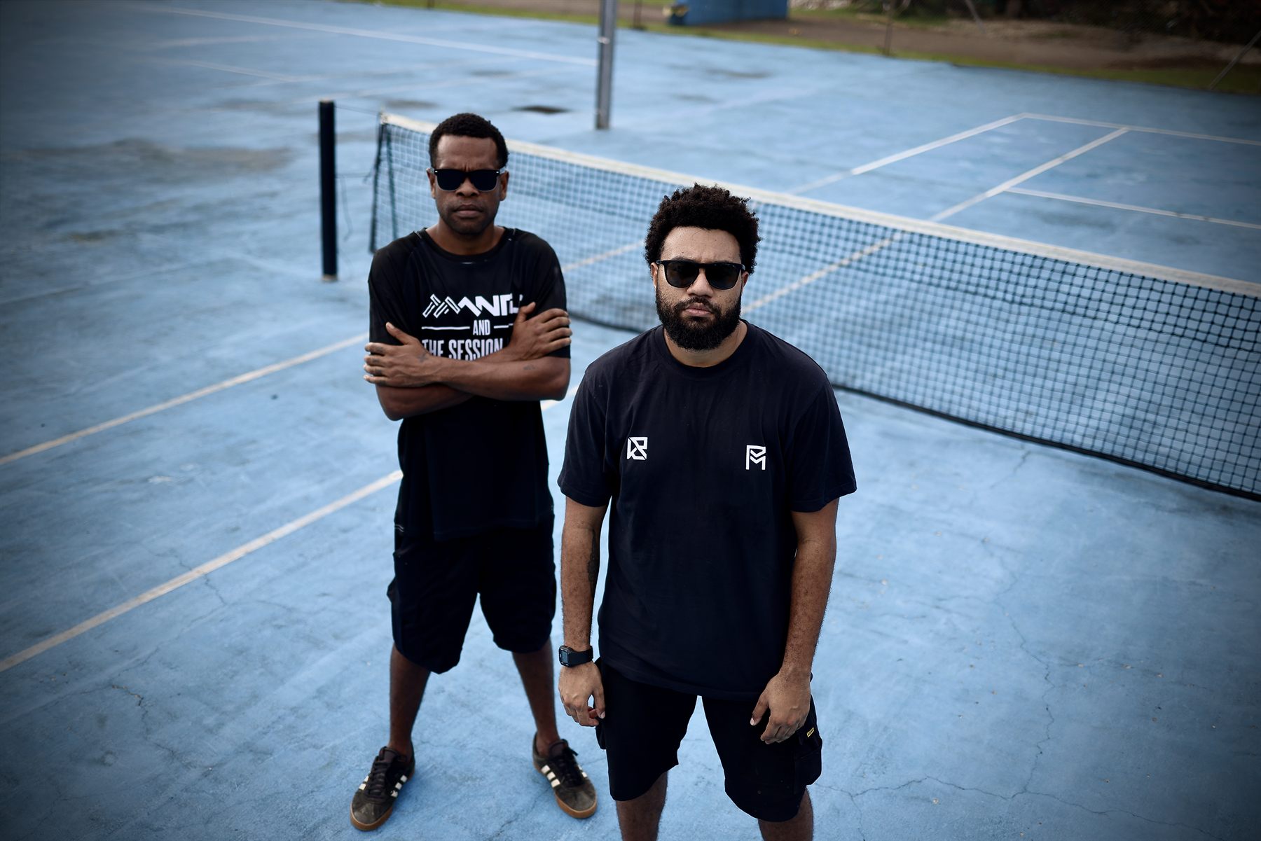 Mantis and his bandmate standing side by side at the net of a weathered blue tennis court, both in black tees and sunglasses, staring straight into the camera.