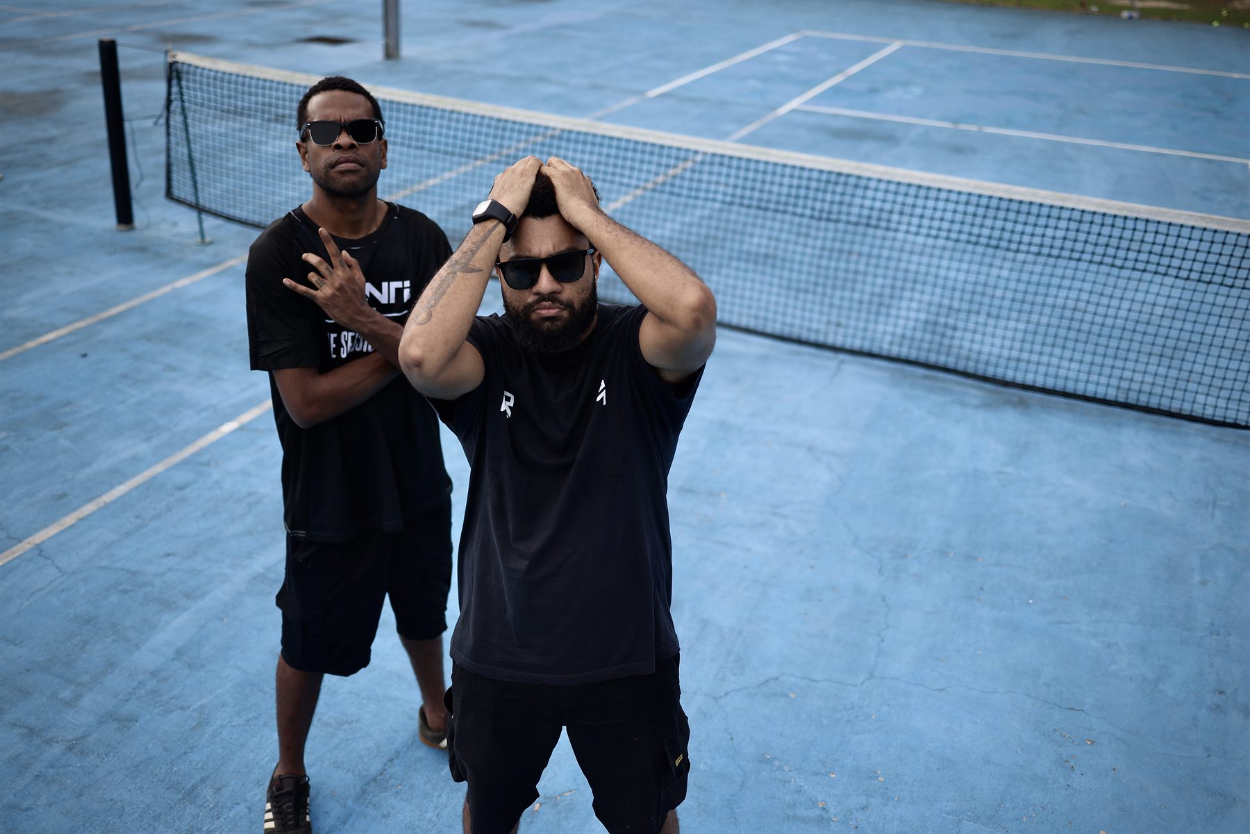 High-angle shot at the net: the bearded artist presses both hands to his head facing the camera while his bandmate poses behind with arms folded.