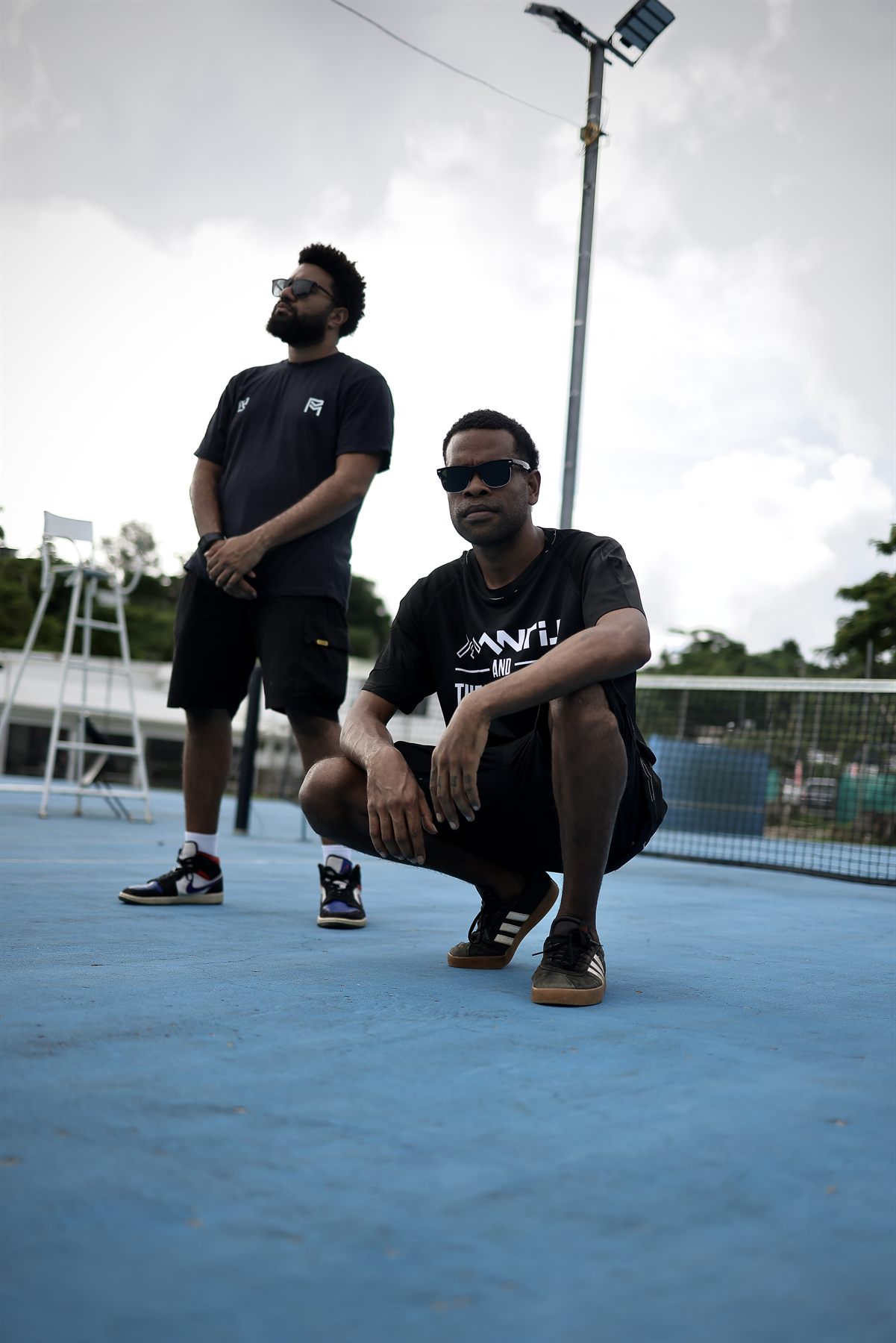 Artist in a Mantis and the Session Band tee crouching on the blue court, forearms on his knees, while his bandmate stands behind gazing skyward beside an umpire chair.