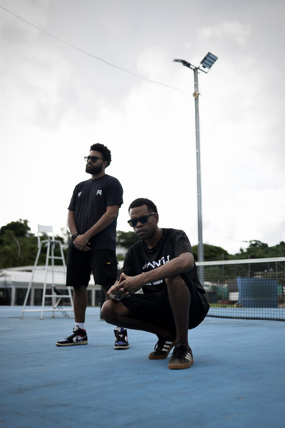Artist in a Mantis and the Session Band tee crouching low with hands clasped, his bandmate standing watch behind him on the blue court under an overcast sky.
